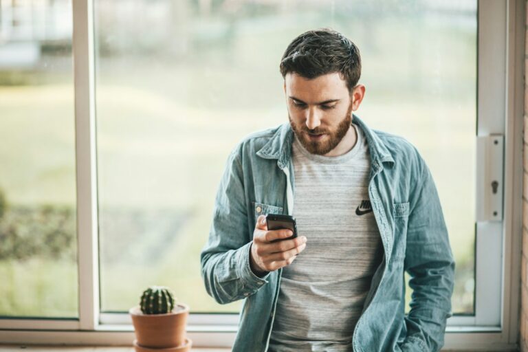 A person holding a phone beside a window, representing the urge to check an ex’s social media after a breakup.
